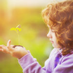 Little girl holding green young plant in spring outdoors. Ecology concept.