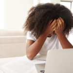 Depressed young teenage black girl with head in hands using laptop computer at home, close up