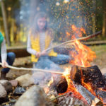Cute little sisters roasting hotdogs on sticks at bonfire. Children having fun at camp fire. Camping with kids in fall forest.
