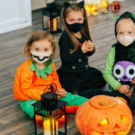 kids in carnival costumes are celebrating Halloween, wearing face masks and playing with pumpkins and candies indoors