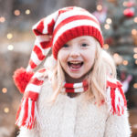 Laughing kid girl 4-5 wearing red knitted hat and scard having fun over Christmas tree outdoors closeup. Looking at camera. Happiness. New Year.