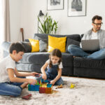 Father using laptop for freelance work sitting on the sofa at home while his two kids playing peacefully together on the floor. Busy single parent is babysitting and working