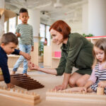 Group of small nursery school children with teacher sitting on floor indoors in classroom, montessori learning.