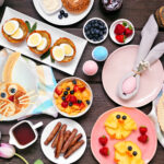 Fun Easter breakfast or brunch table scene. Top down view on a dark wood background. Bunny pancake, egg nests, chick fruit and assorted spring food items.