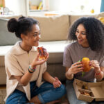 Portrait of young sisters indoors at home, eating burgers.