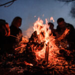 Young and cheerful friends sitting and fry marshmallows near bonfire at night