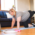 Online fitness training for overweight woman exercising at home, looking at laptop. Workout in living room.