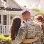 A male veteran american soldier in military uniform hugs his wife and little daughter while standing in front of their house. The concept of a emotional military happy homecoming. Sunny day