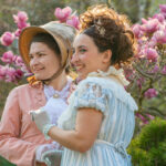 Two women in Regency-era outfits in a garden during spring.
