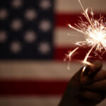 Hand holding lit sparkler in front of the American Flag for 4th of July celebration